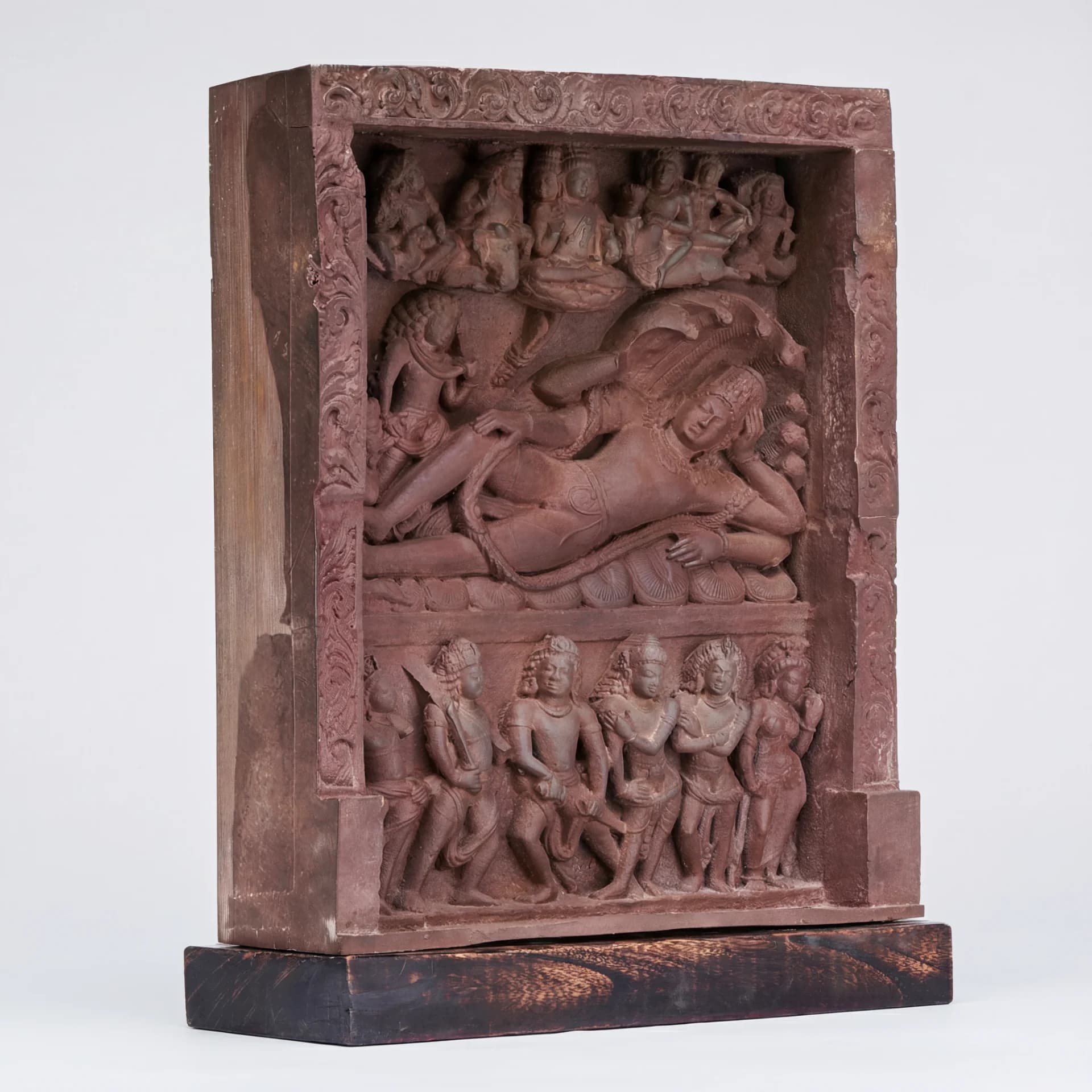 A three-quarter view of the Anantasayana Vishnu Sculpture, a terracotta-style replica featuring intricate relief carvings of deities on a dark wooden base.