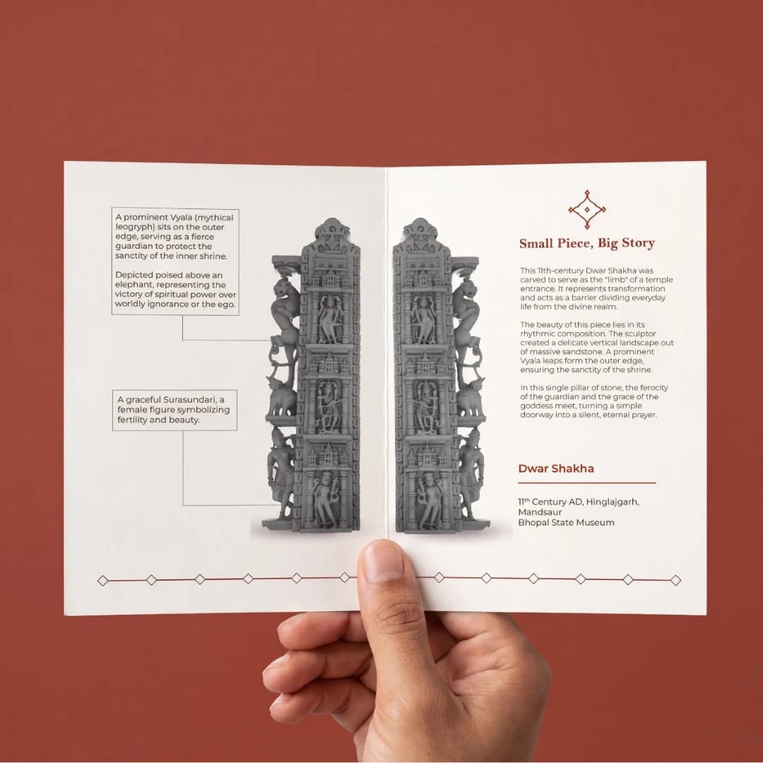 A hand holds an open brochure displaying the Dwara Shakha Sculpture, a stone composite replica featuring detailed carvings of celestial figures.
