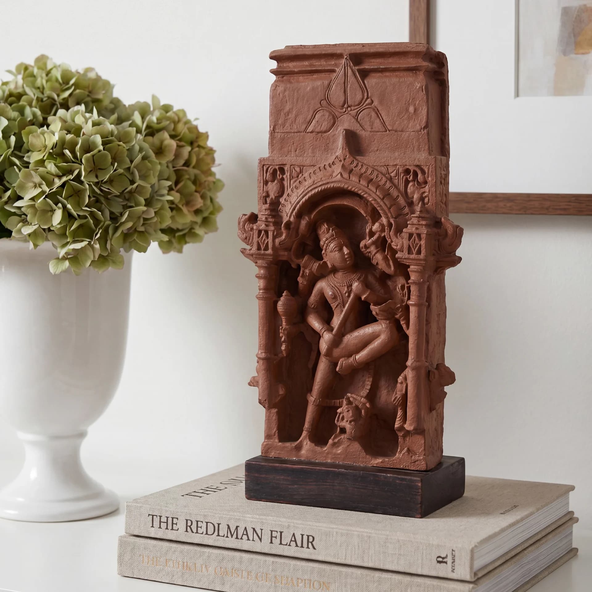 The Gajasura Vadha Sculpture displayed on stacked books, featuring intricate terracotta-style relief carvings of Shiva in a temple archway.