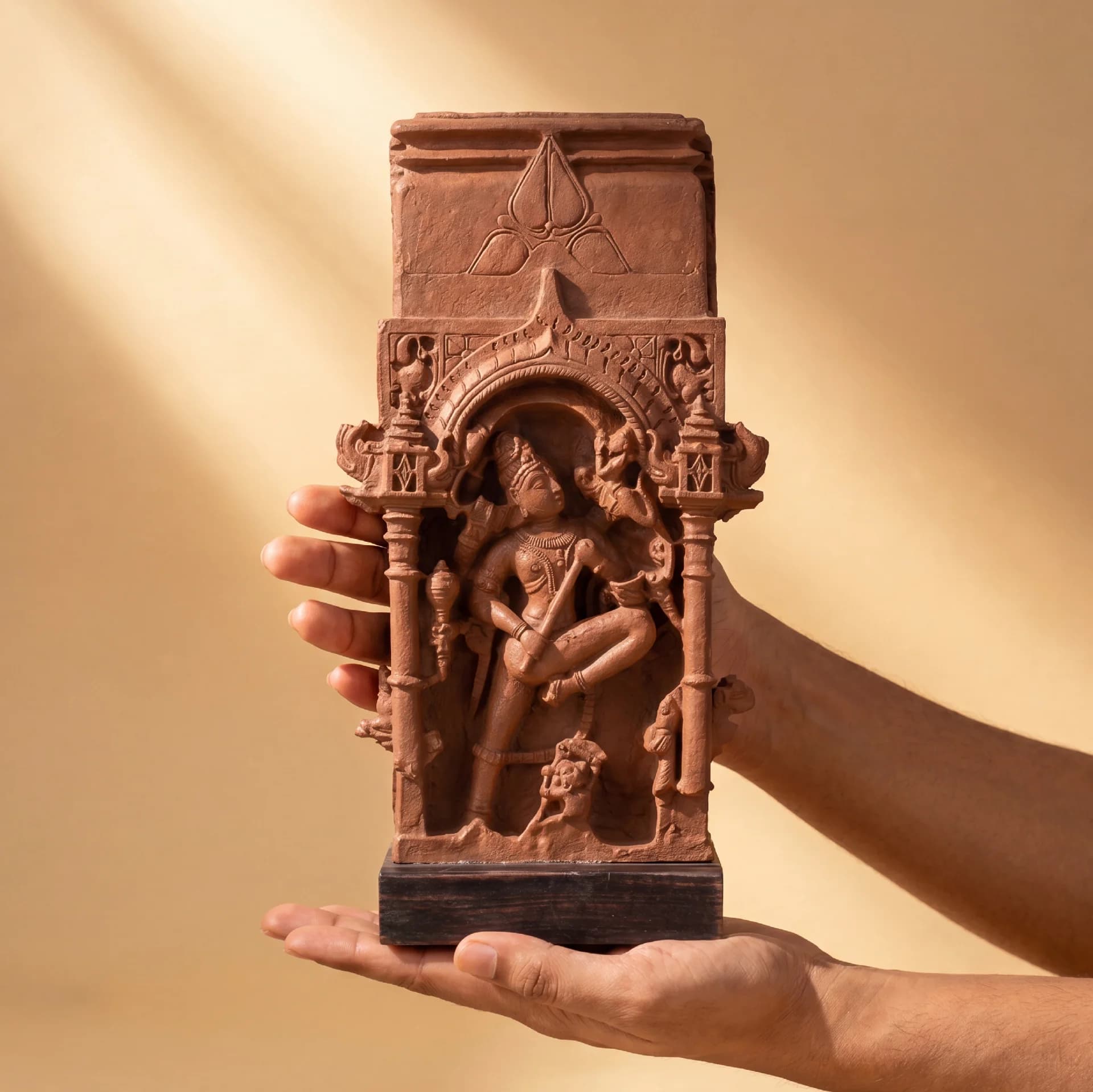 Hands hold a terracotta-style Gajasura Vadha Sculpture, showcasing the intricate 10th-century temple relief carving against a soft background.