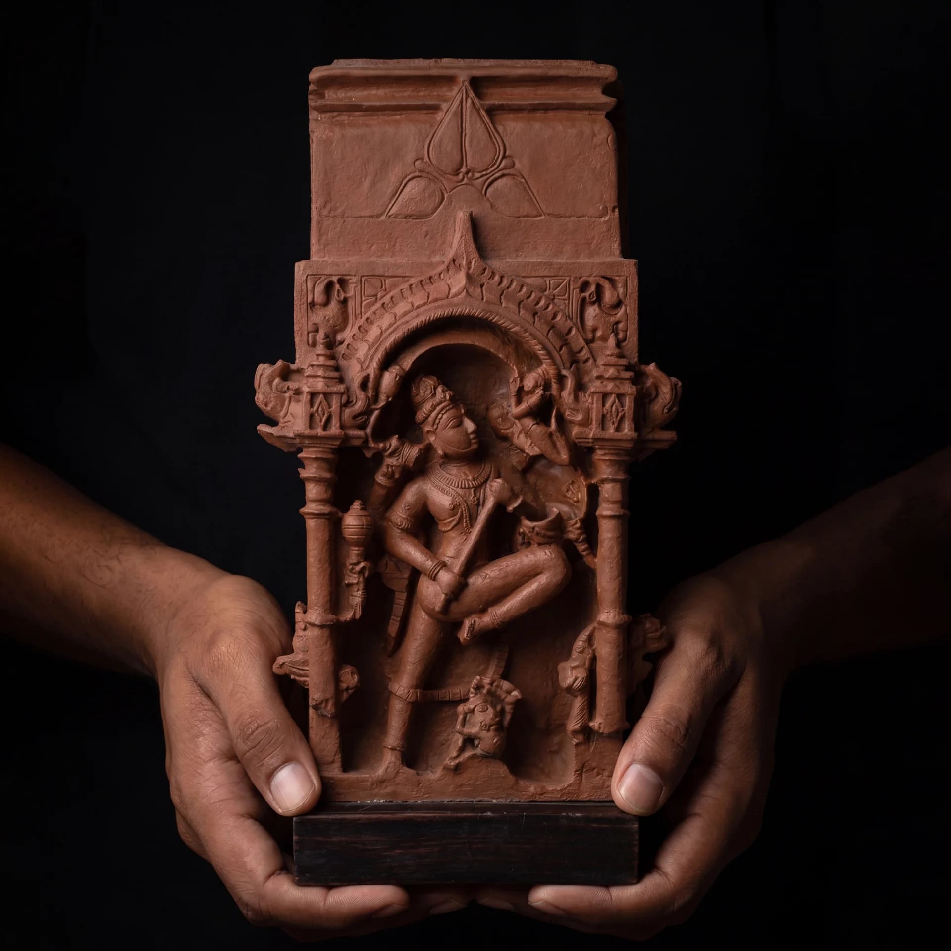 Close-up of hands holding the terracotta-style Gajasura Vadha Sculpture, showcasing the intricate 10th-century temple relief carving details.
