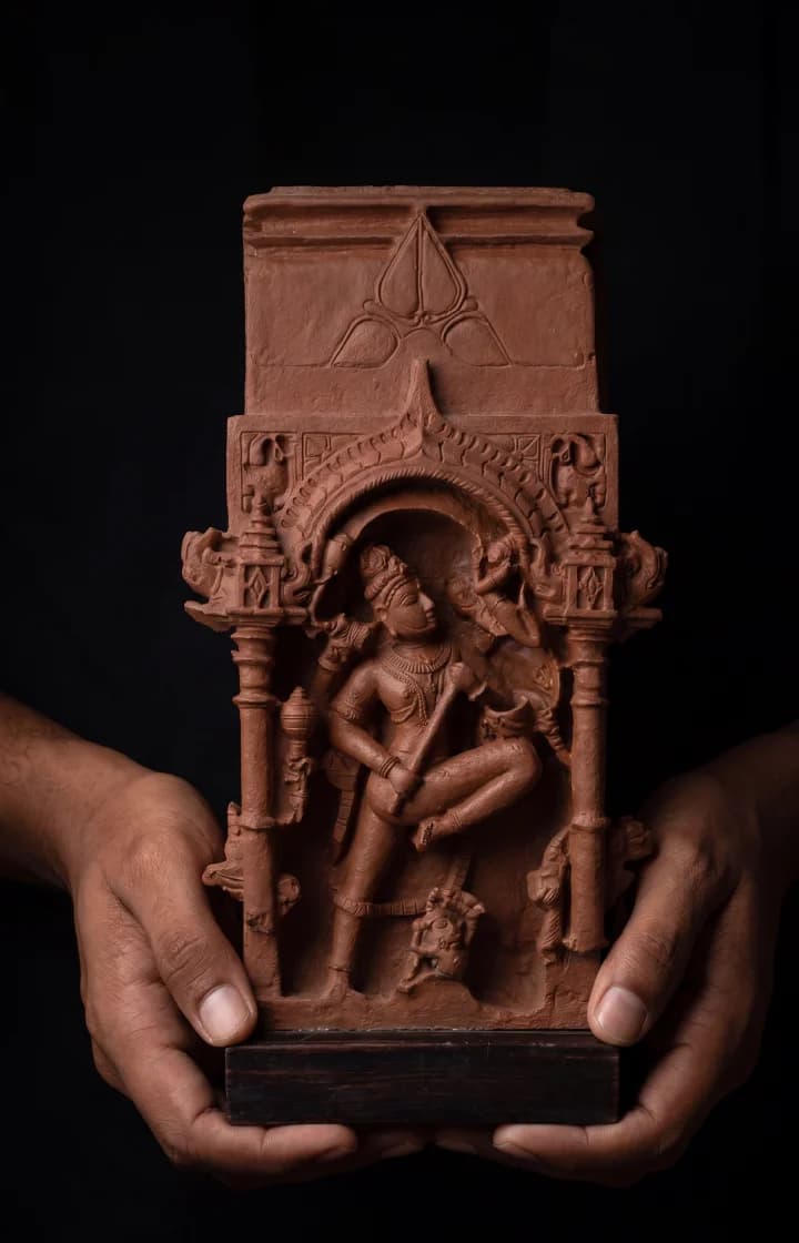 Hands holding a terracotta-style Gajasura Vadha Sculpture, showcasing the intricate temple relief carving of Shiva in a black background.