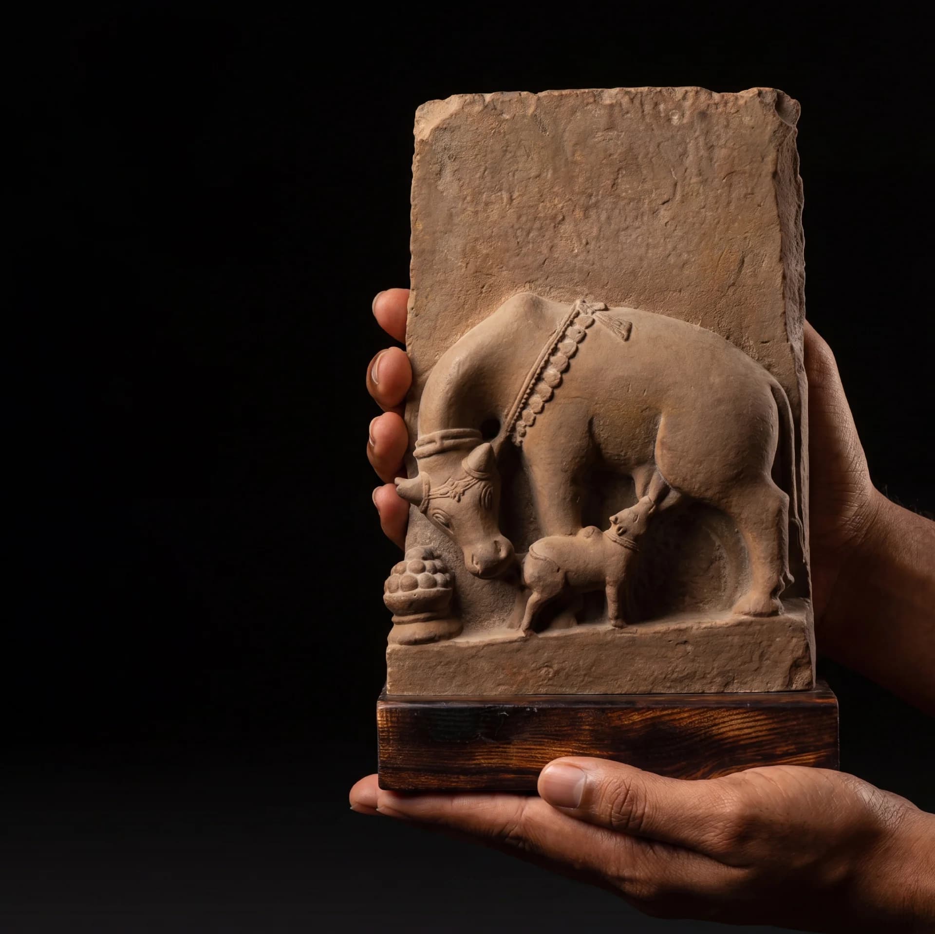A pair of hands holds a hand-finished stone composite Kamdhenu Sculpture depicting a divine cow nursing her calf.
