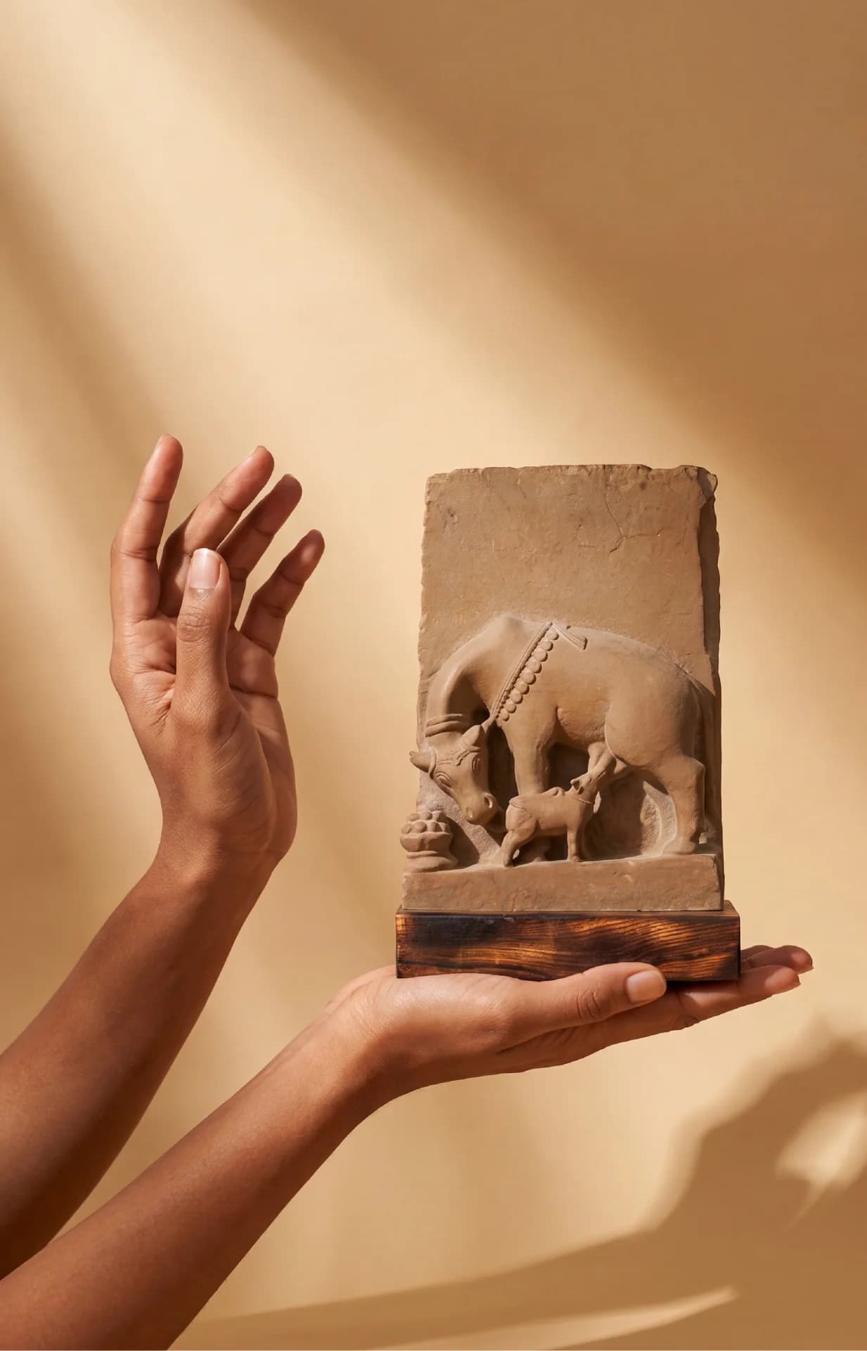 A person's hands hold a stone composite Kamdhenu Sculpture, depicting the divine cow and calf in a museum-replica style.