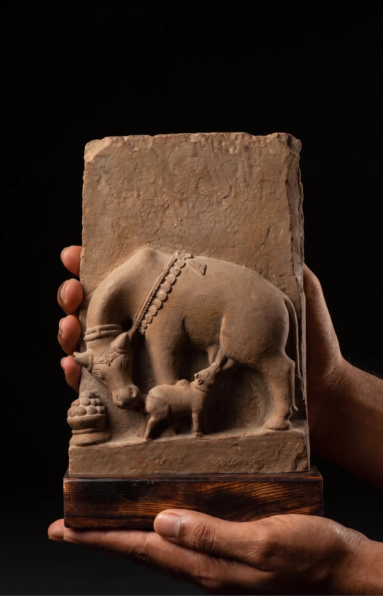 A pair of hands holds a stone composite Kamdhenu Sculpture, a detailed museum replica depicting a cow nursing her calf.