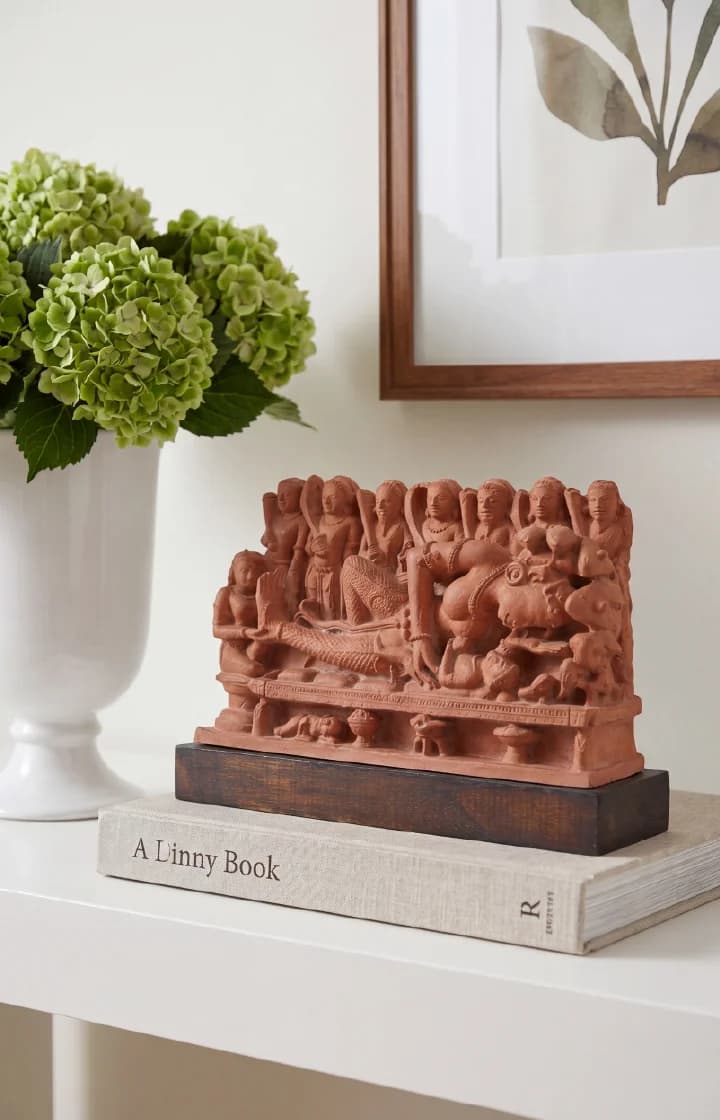 A terracotta-style Krishna's Birth Sculpture displayed on a book, showing intricate relief carvings of figures on a wooden base.