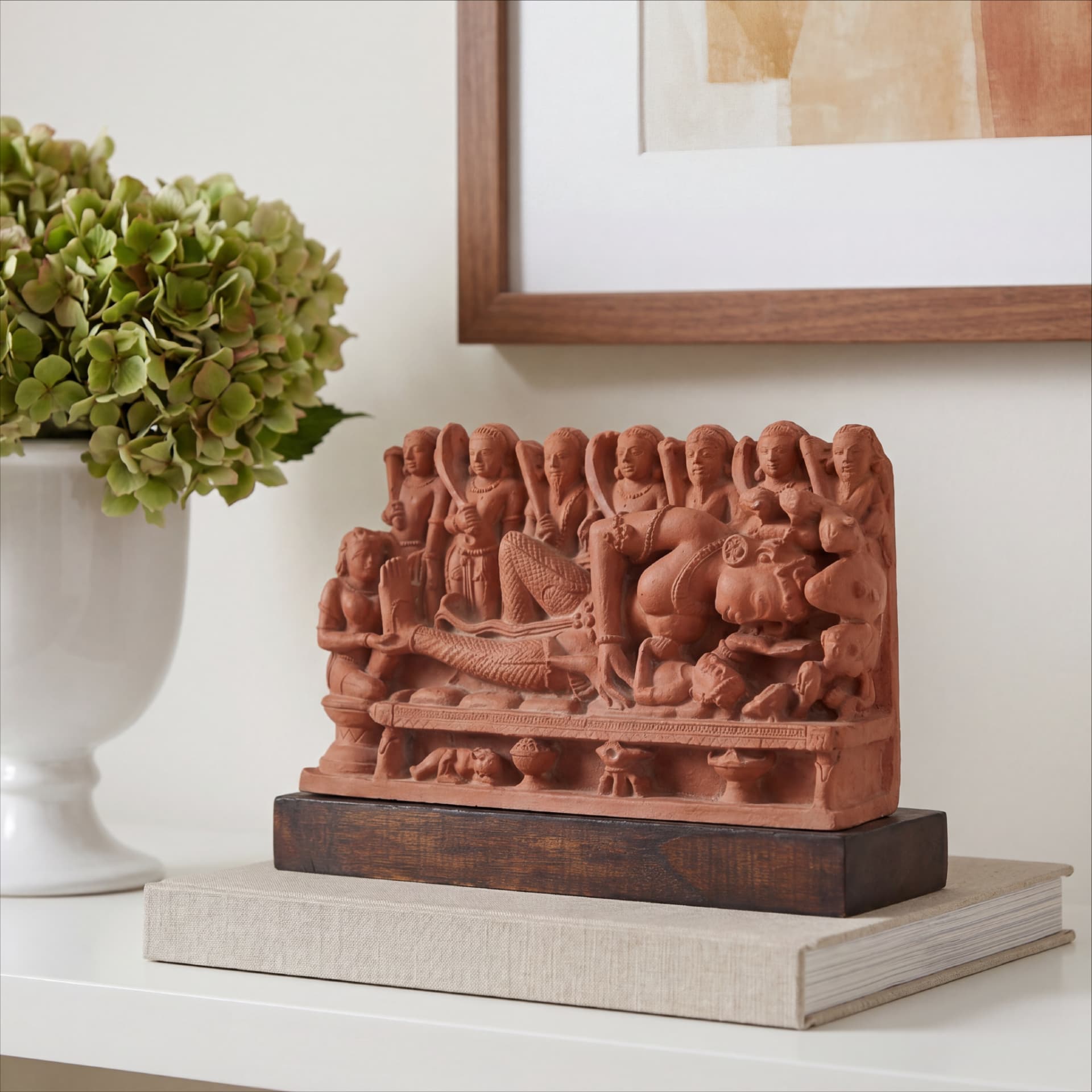 A terracotta Krishna's Birth Sculpture displayed on a book next to a white vase with green hydrangeas.