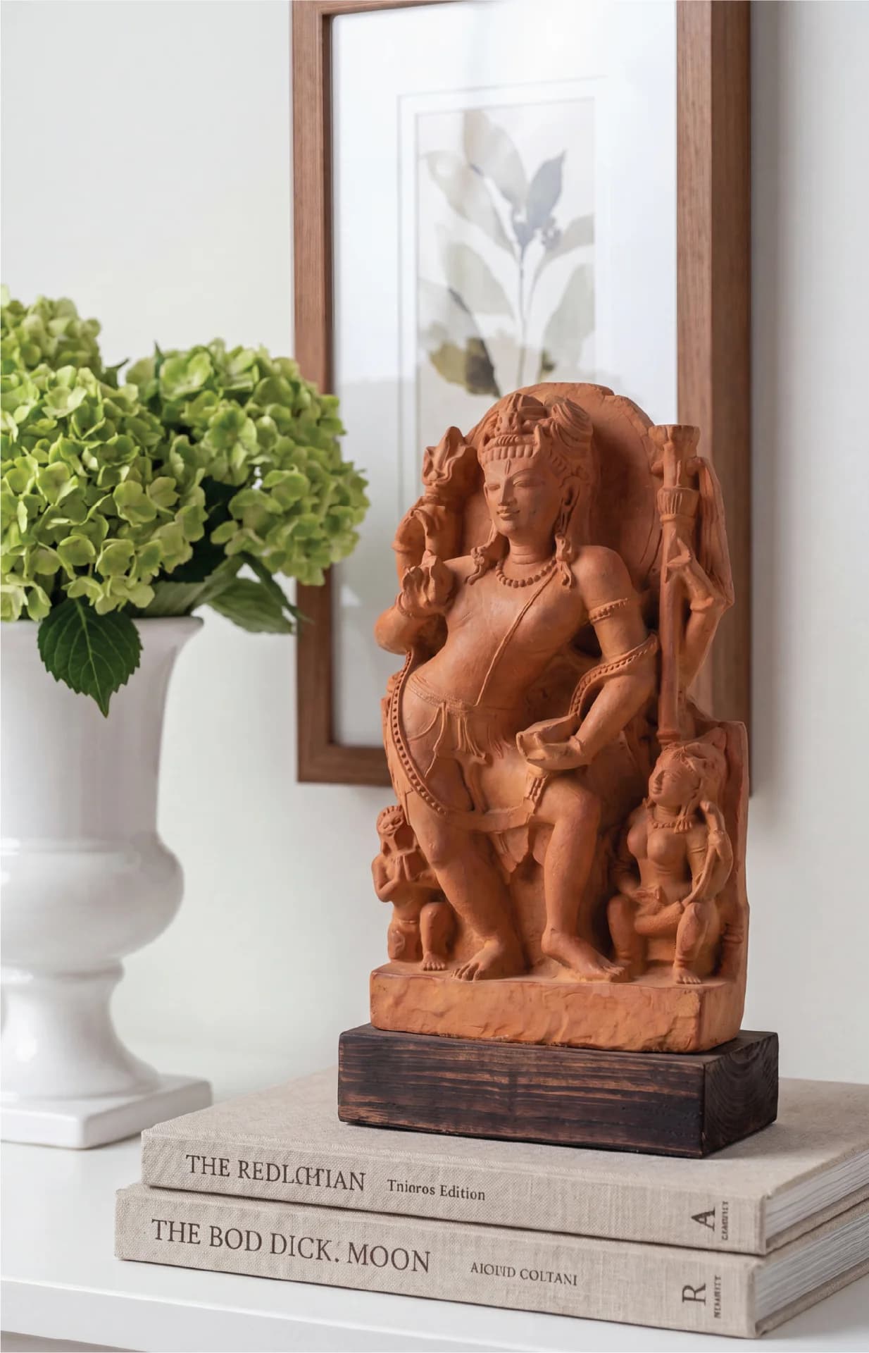 A stone composite Natesh Shiva Sculpture displayed on stacked books next to a white vase with green flowers.