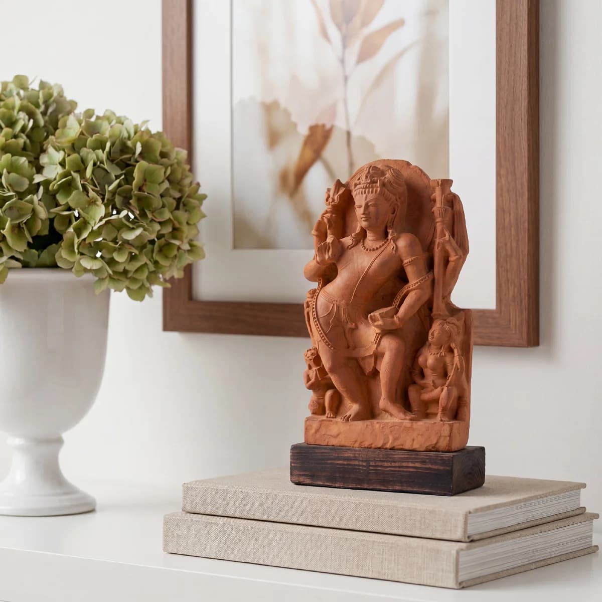 A stone composite Natesh Shiva Sculpture displayed on stacked books next to a white vase with green flowers.