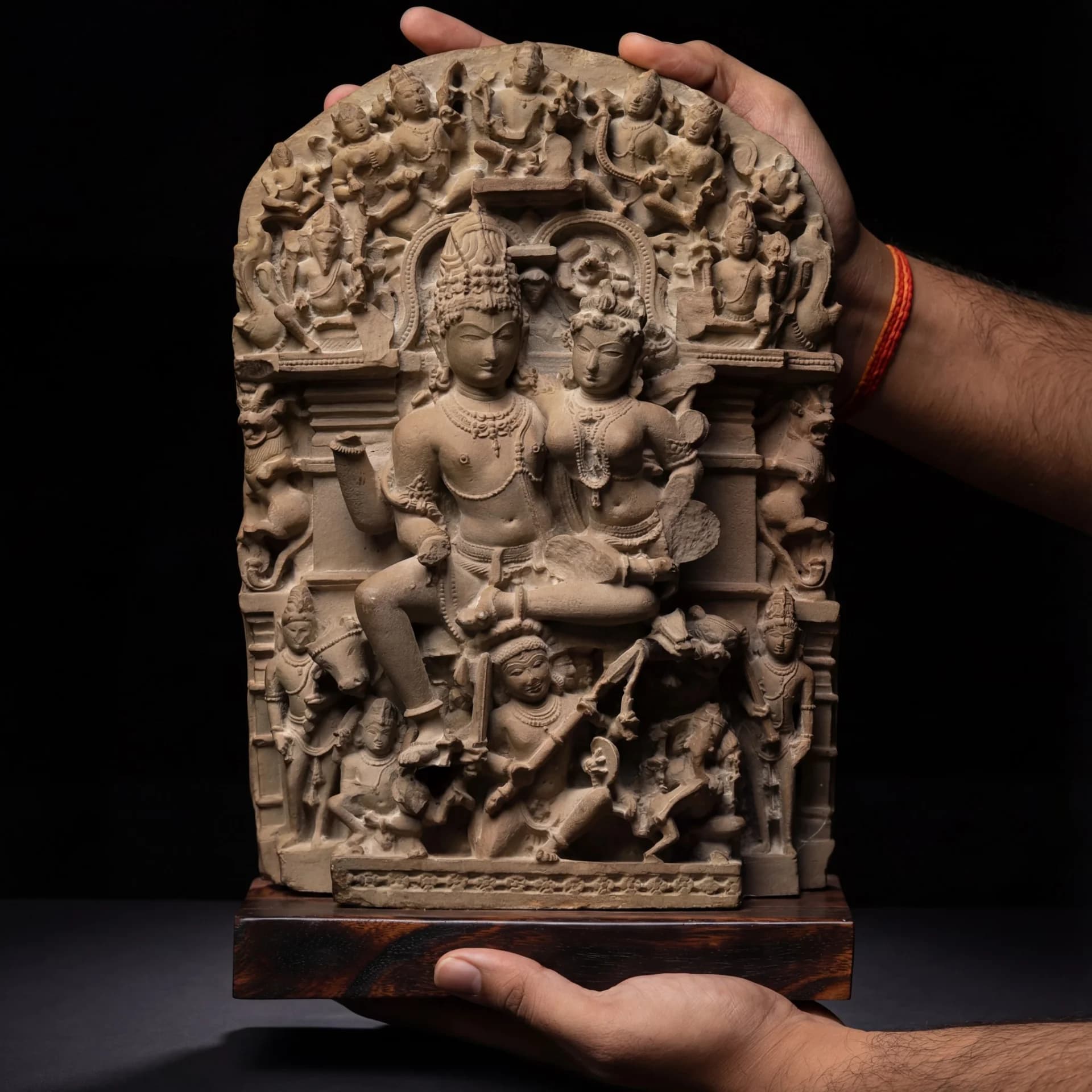 Hands holding a stone composite Ravananugraha Sculpture replica featuring intricate carvings of Shiva and Parvati against a black background.