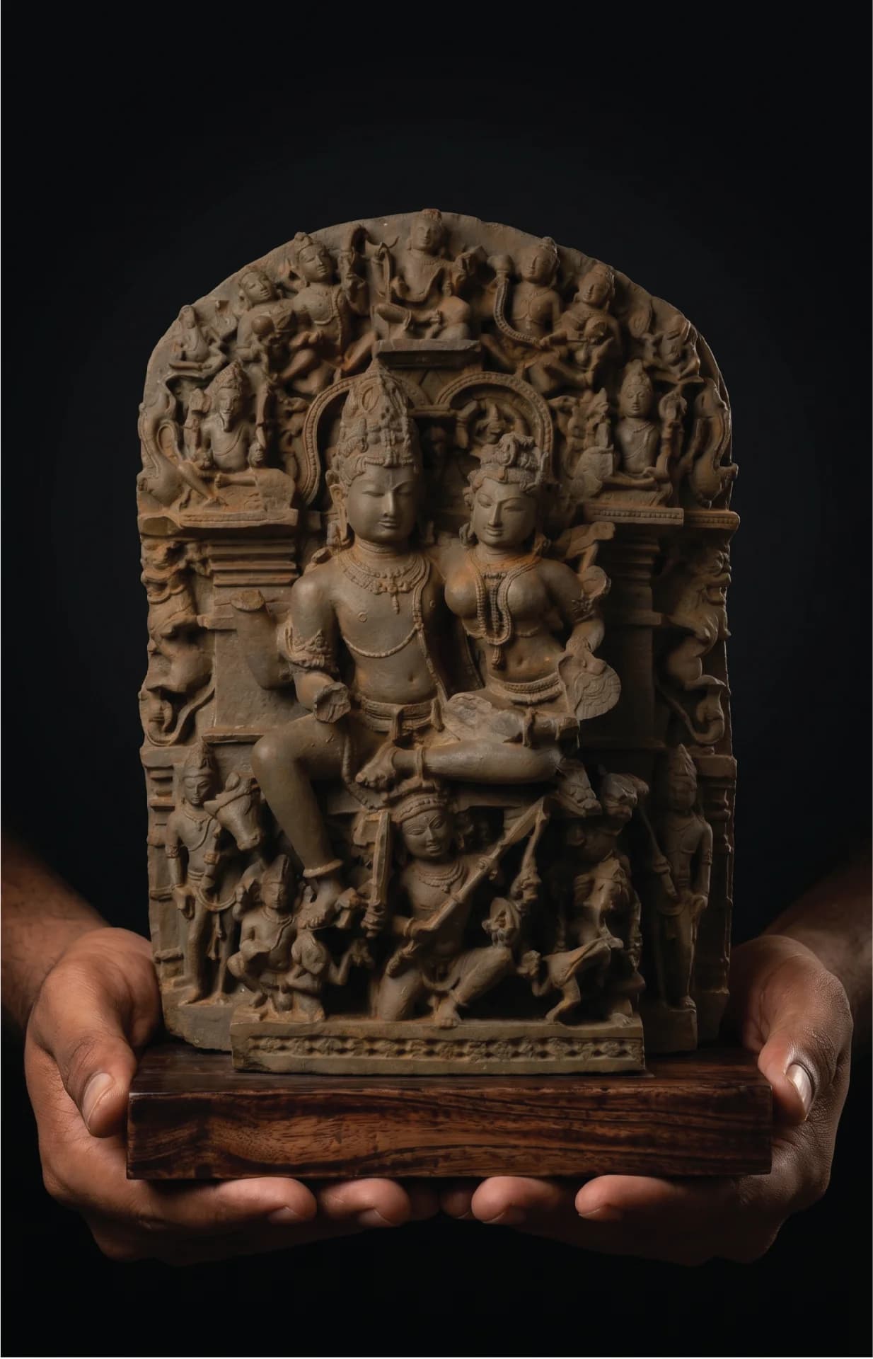 A pair of hands holds a stone composite Ravananugraha Sculpture, showcasing intricate 11th-century temple art on a teak base.