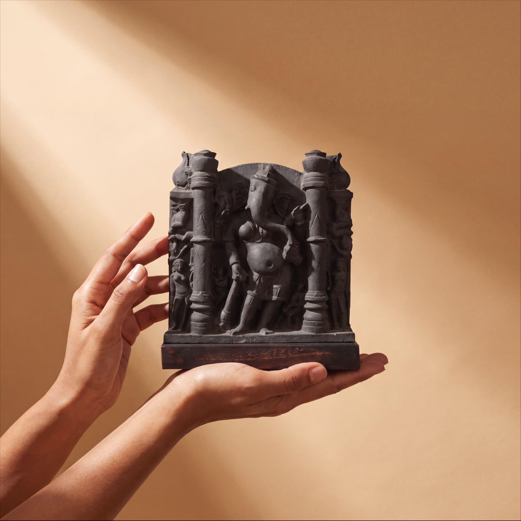 A pair of hands gently holds a dark stone Vinayaki Sculpture, showcasing the intricate 11th-century temple art replica details.