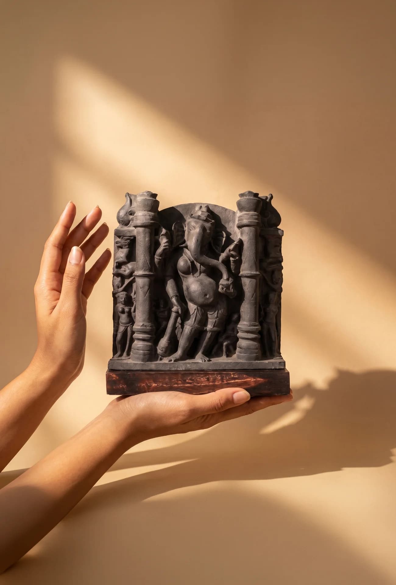 A pair of hands gently holds the Vinayaki Sculpture, a dark stone-finish museum replica featuring intricate 11th-century temple iconography.