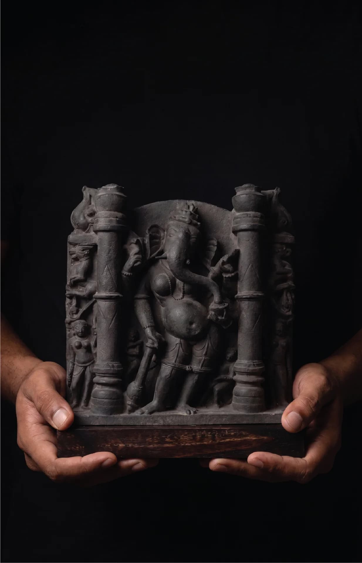 A pair of hands holds a dark stone Vinayaki Sculpture, showcasing the intricate 11th-century medieval Indian temple art style.