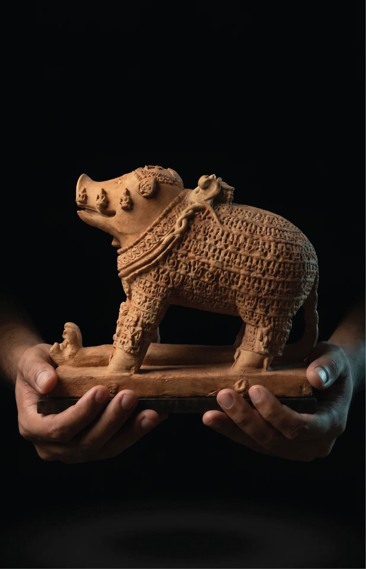 Hands holding an Adi Varaha Sculpture, a detailed terracotta-style museum replica featuring intricate carvings of divinities across the boar's body.