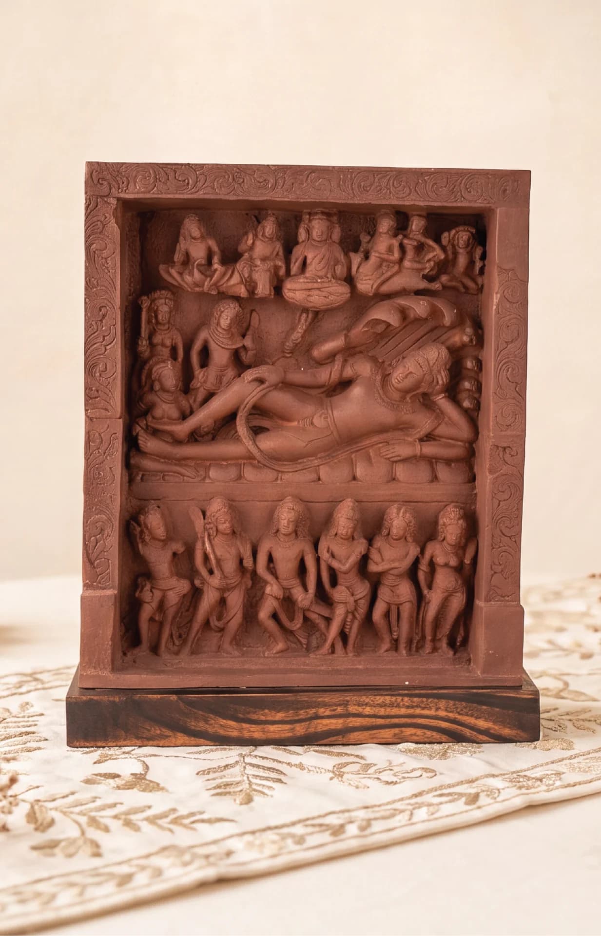 A front-facing view of the Anantasayana Vishnu Sculpture, a terracotta-style replica featuring intricate relief carvings of deities on a wooden base.