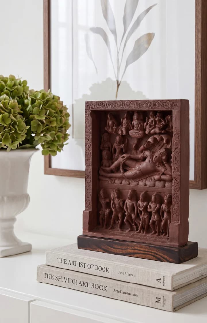 An Anantasayana Vishnu Sculpture museum replica displayed on a stack of books next to a white vase with greenery.