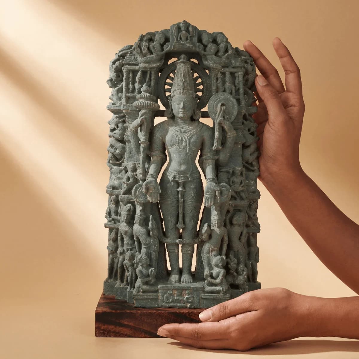 A pair of hands gently holds the stone composite Chaturbhuj Vishnu Sculpture, highlighting the intricate carvings of the deity.