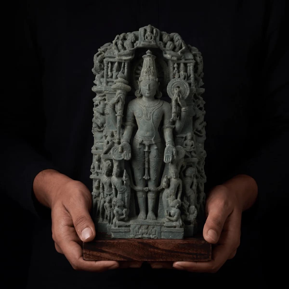 A pair of hands holds a stone composite Chaturbhuj Vishnu Sculpture, showcasing the intricate standing deity and carved parikara details.