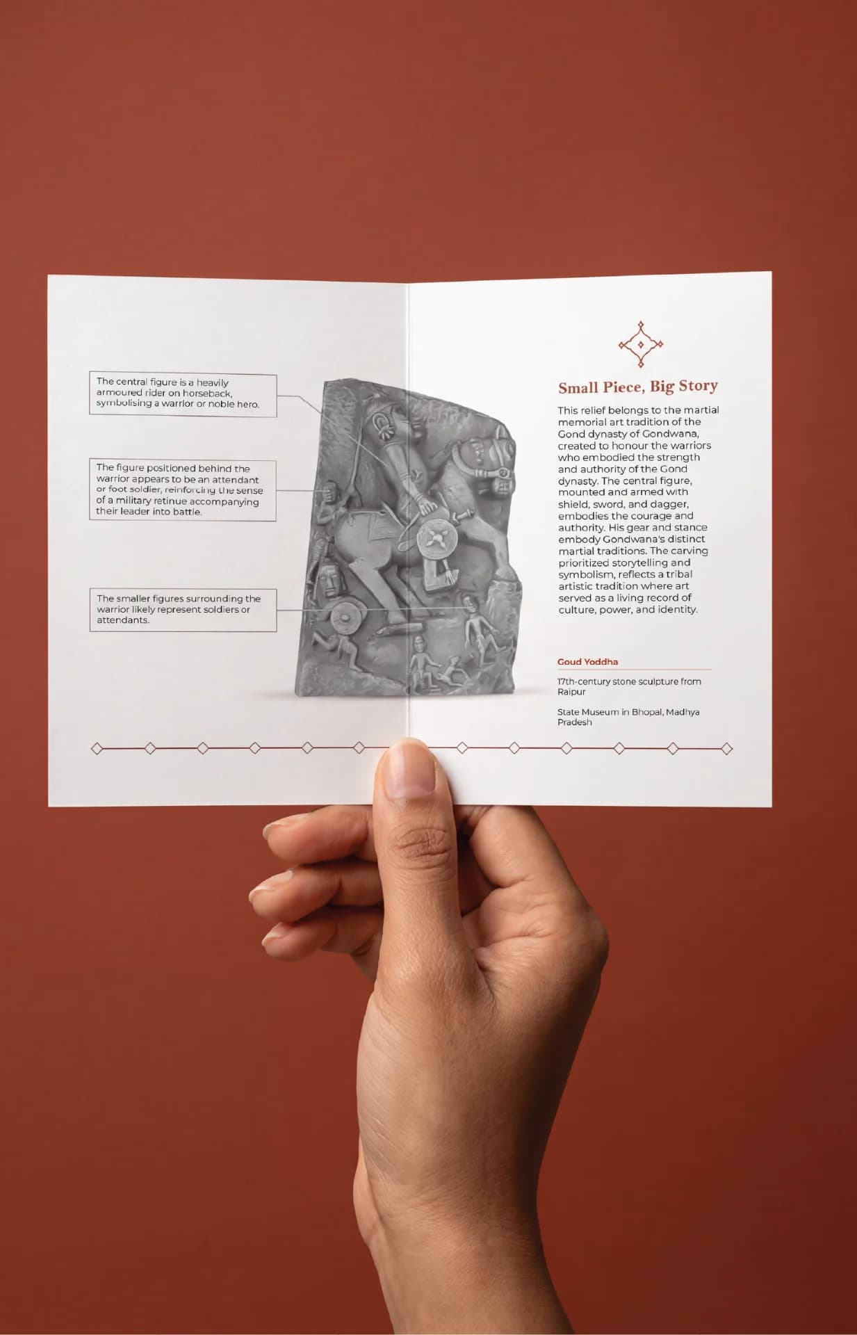 A hand holds an open brochure featuring the Goud Yoddha stone sculpture with detailed annotations explaining the historical relief.