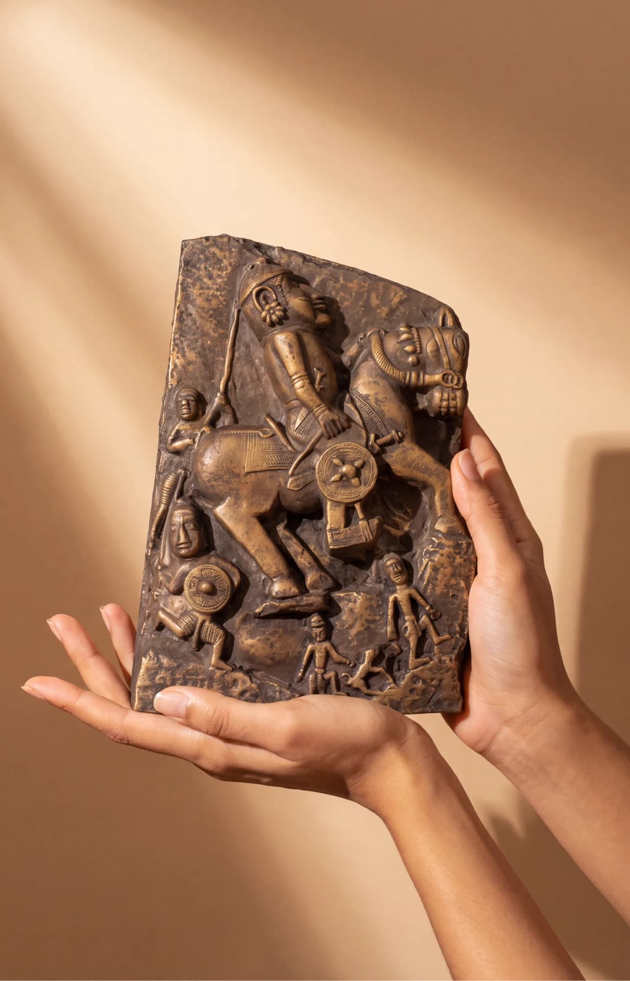 Two hands hold a Goud Yoddha stone-style relief sculpture depicting a mounted warrior and smaller figures in relief.