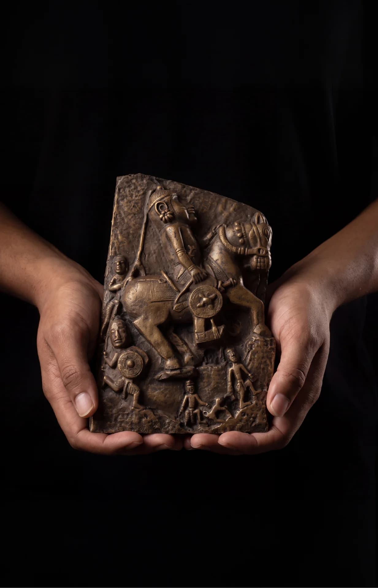 A pair of hands holds the Goud Yoddha, a detailed stone-finish relief sculpture depicting a historic Gond warrior on horseback.