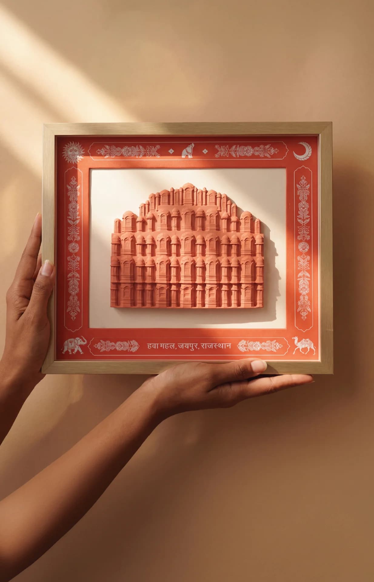 A pair of hands holds a framed Hawa Mahal Facade Sculpture featuring intricate terracotta-style relief work on a decorative mat.