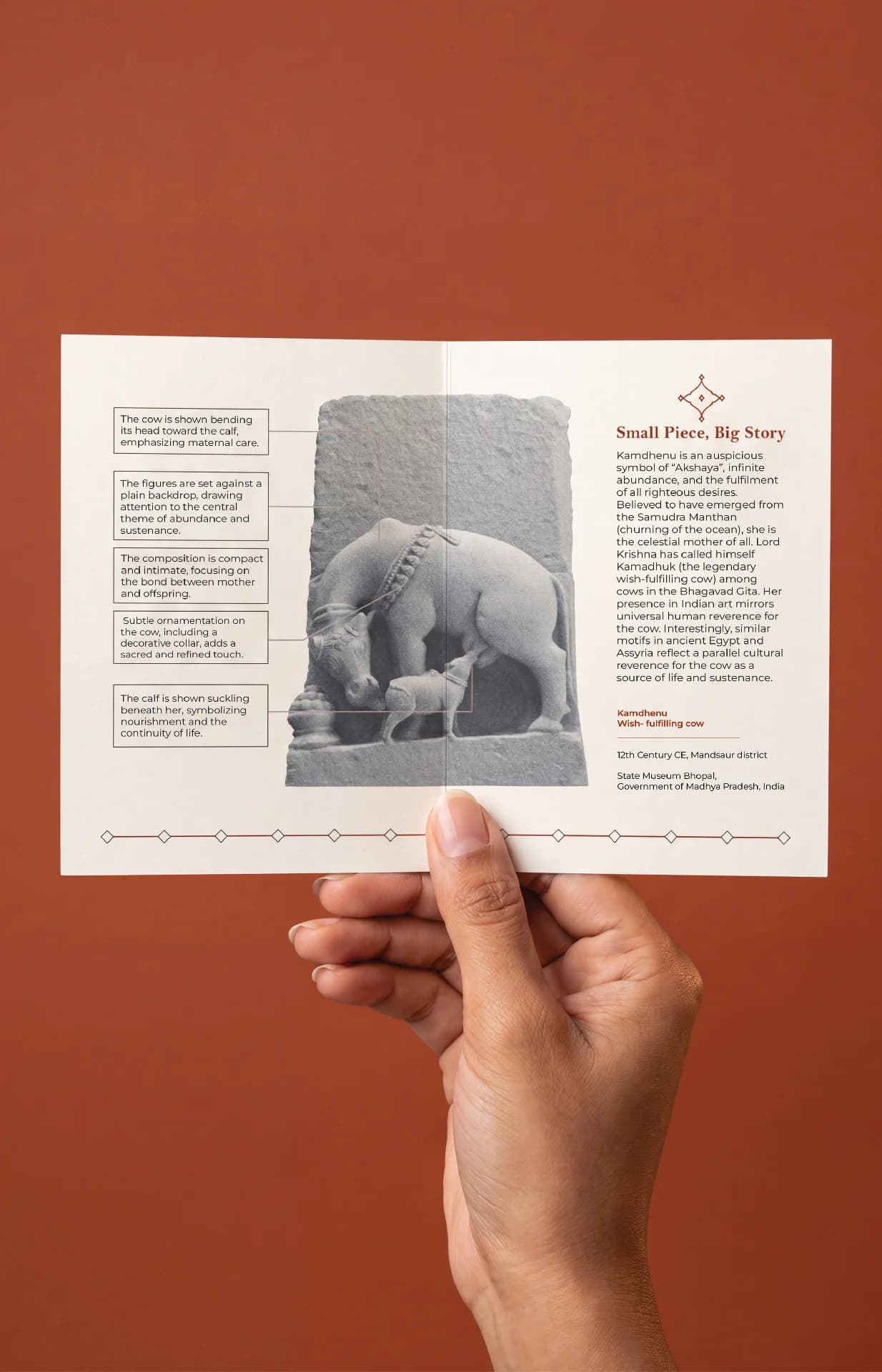 A hand holds an informational card featuring the Kamdhenu Sculpture, a stone composite museum replica depicting a divine cow nursing her calf.