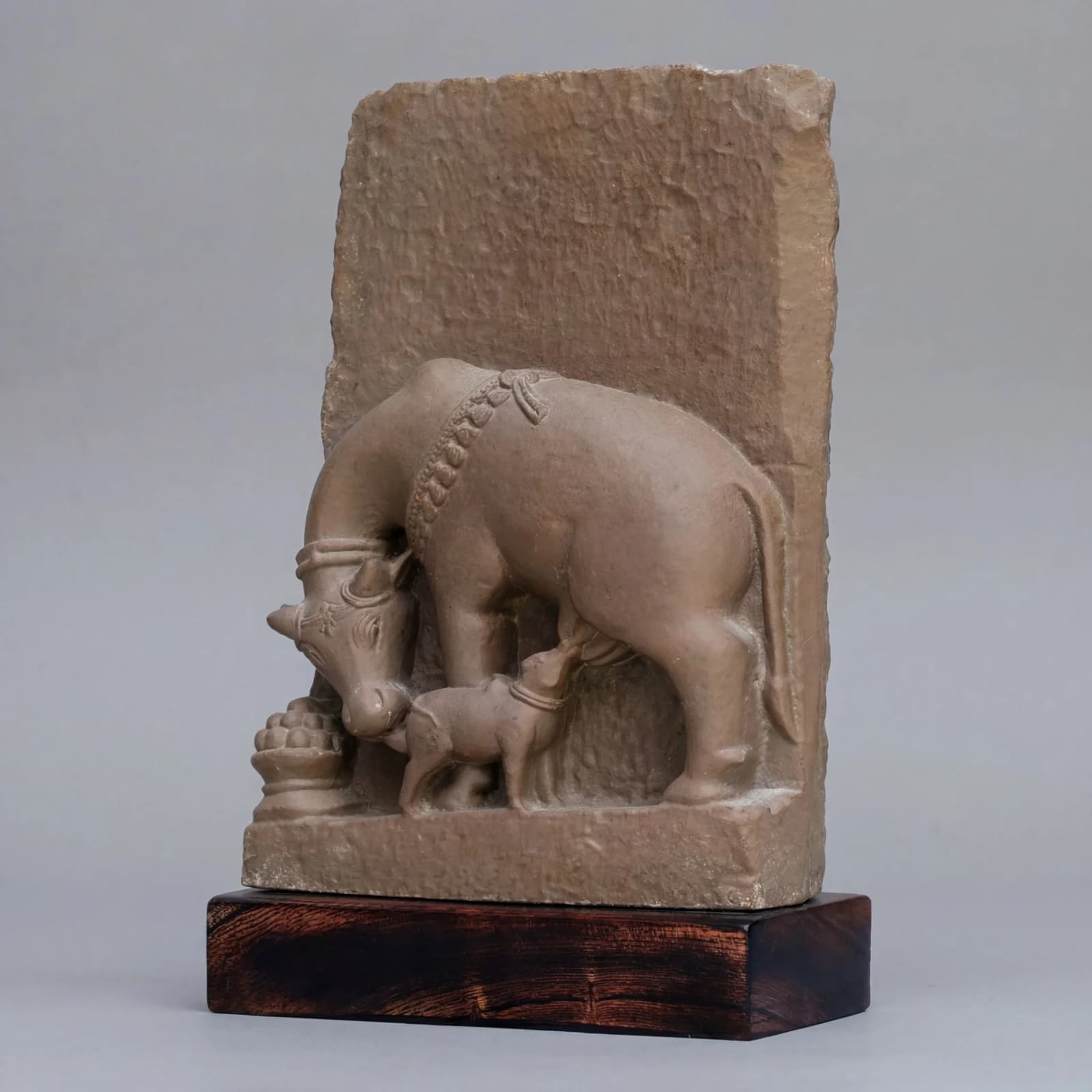 A stone composite Kamdhenu Sculpture showing a mother cow nursing her calf, displayed on a dark wooden base.