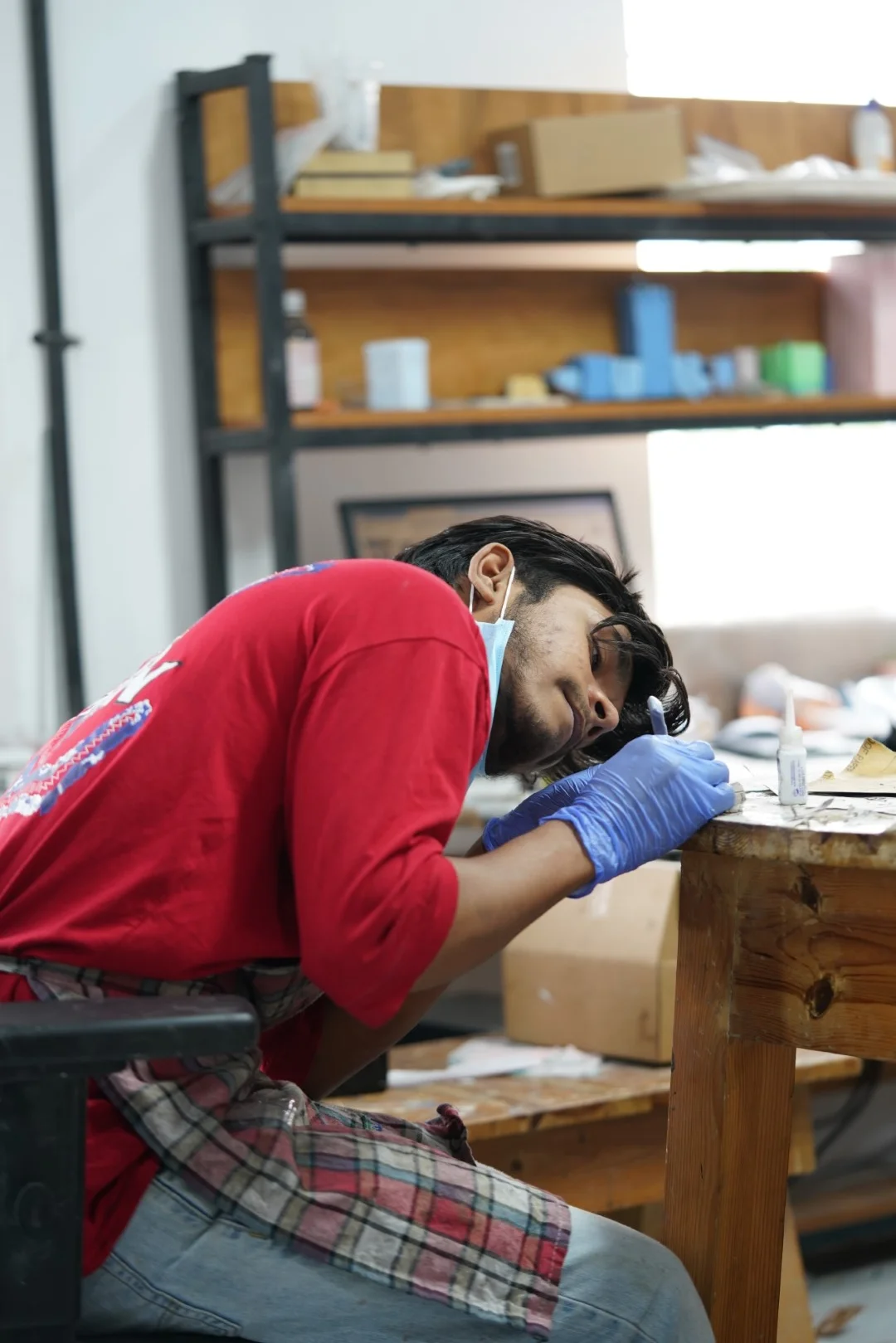 An artisan wearing blue gloves and a red shirt meticulously crafting a museum-replica artifact in a workshop studio.