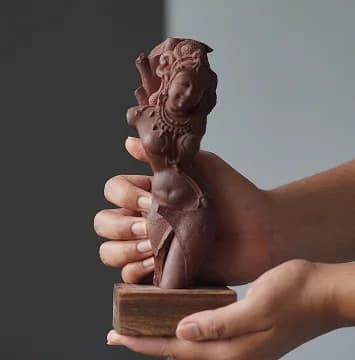 A pair of hands gently cradles a terracotta apsara sculpture, highlighting the intricate carved details and smooth clay texture.
