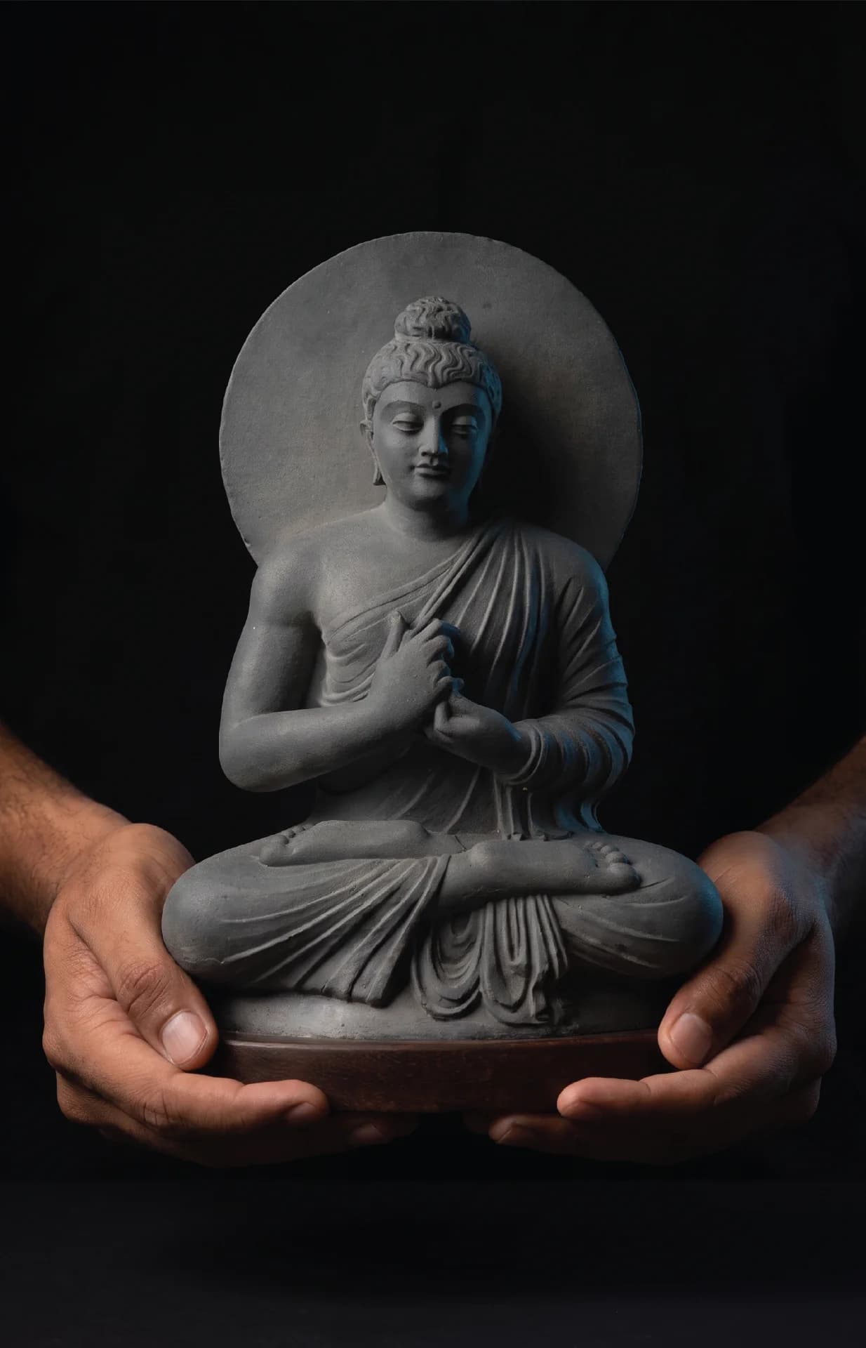 A pair of hands holds a stone composite Preaching Buddha statue, showcasing the intricate Gandhara style against a black background.