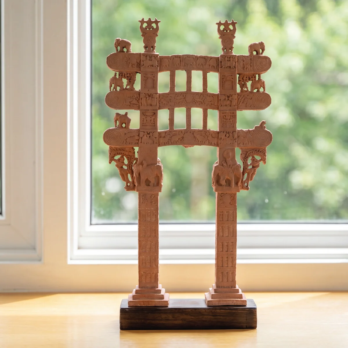 A Sanchi Torana Gateway Miniature featuring intricate terracotta-style carvings of elephants and figures, displayed against a bright window.