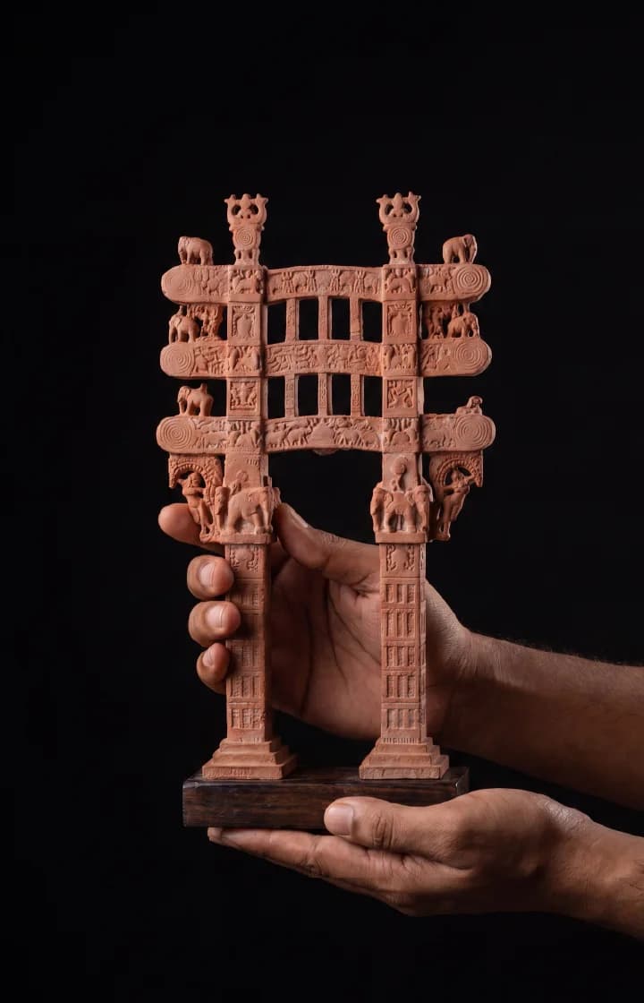 A pair of hands holds a terracotta Sanchi Torana Gateway Miniature, showcasing intricate carvings against a dark background.