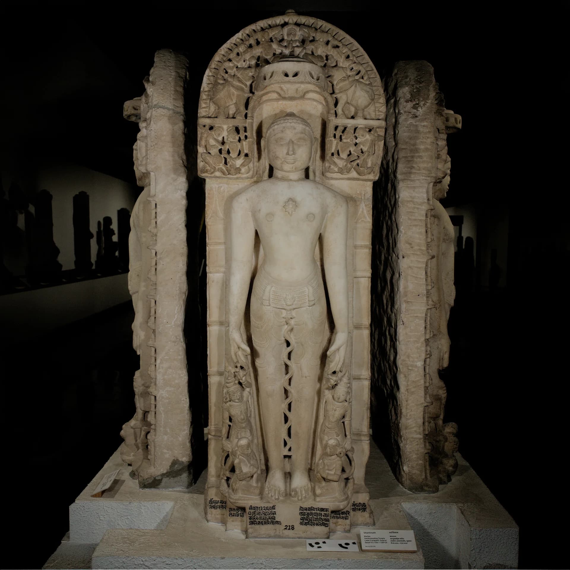 A front-facing view of the Shantinath Sculpture, a detailed marble museum replica featuring intricate carvings and a lattice canopy.