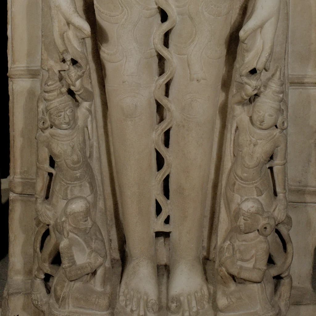 Close-up of the Shantinath Sculpture marble replica showing intricate carvings of flanking figures at the base of legs.