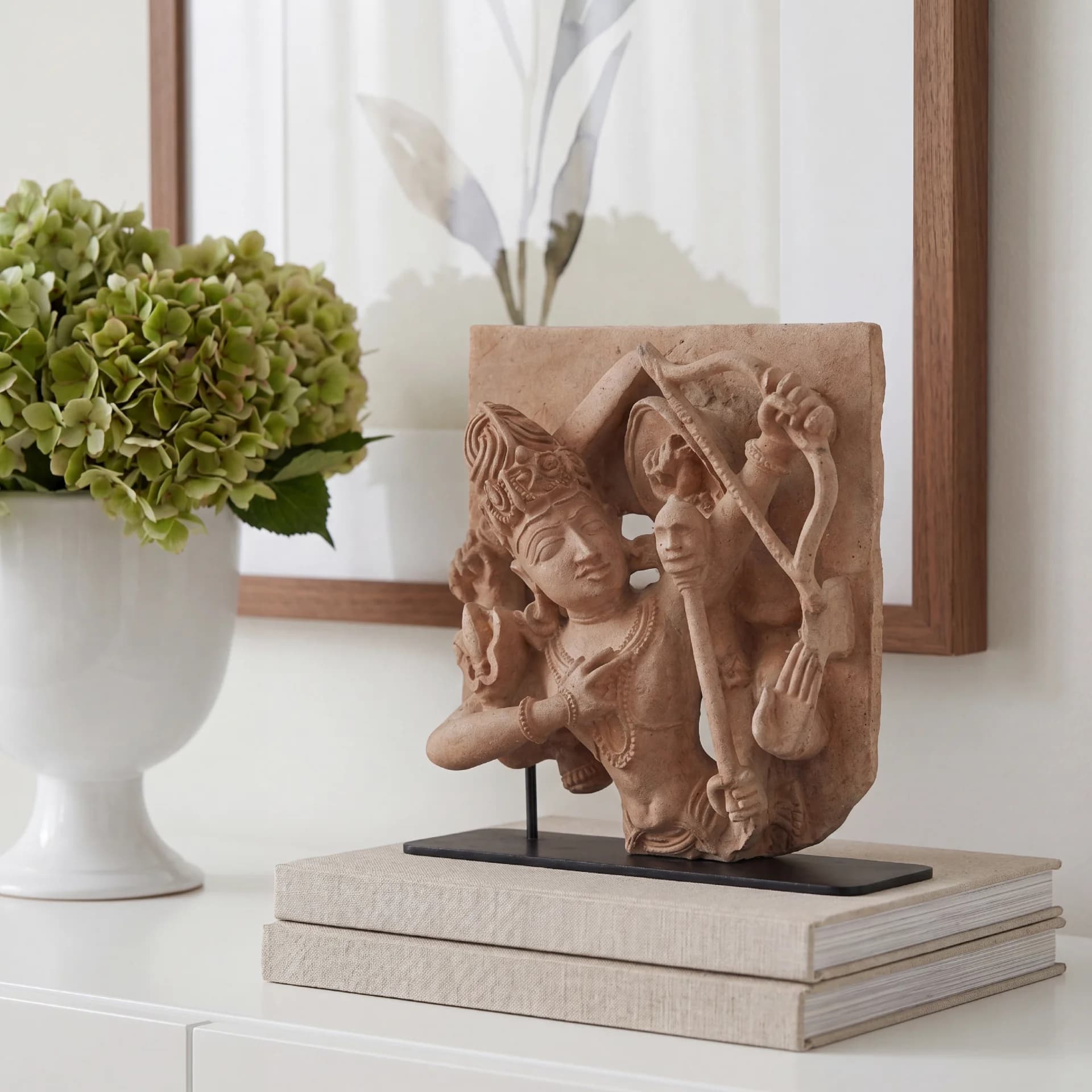 A stone composite Tripurantaka Shiva sculpture displayed on stacked books next to a white vase with green flowers.