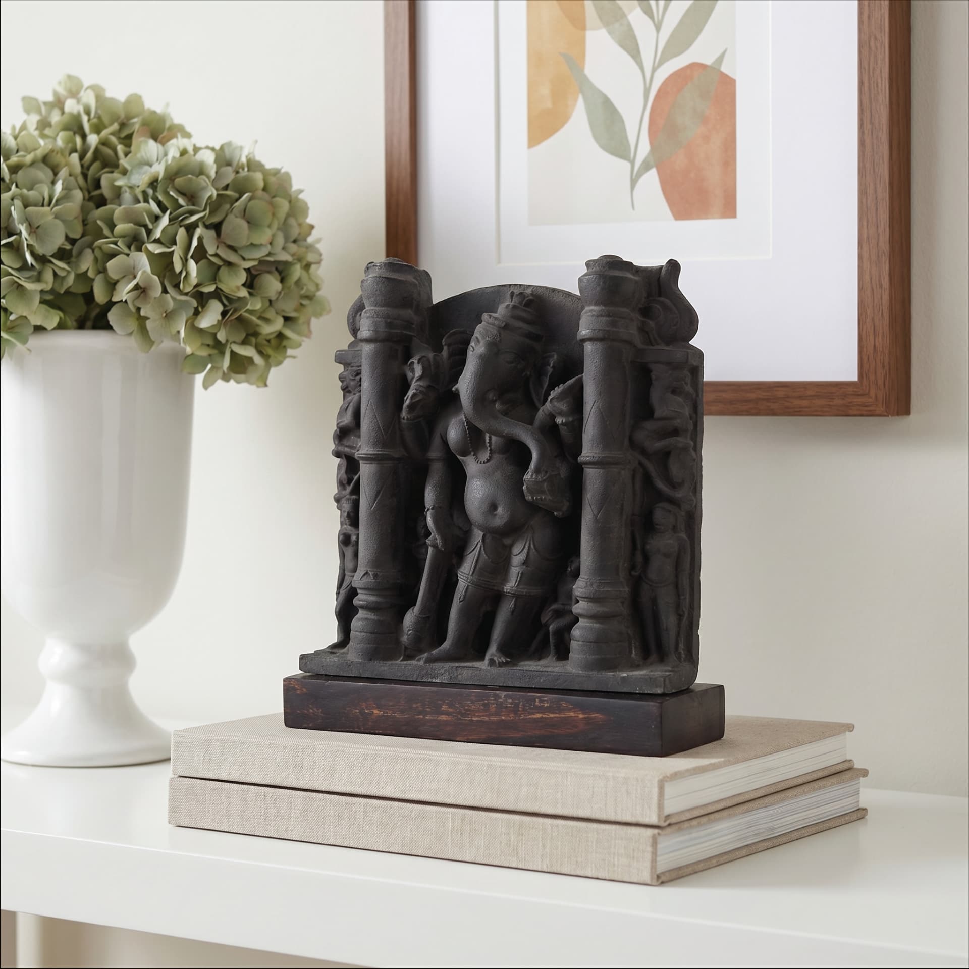 A dark stone-finish Vinayaki Sculpture displayed on a stack of books next to a white vase with green flowers.