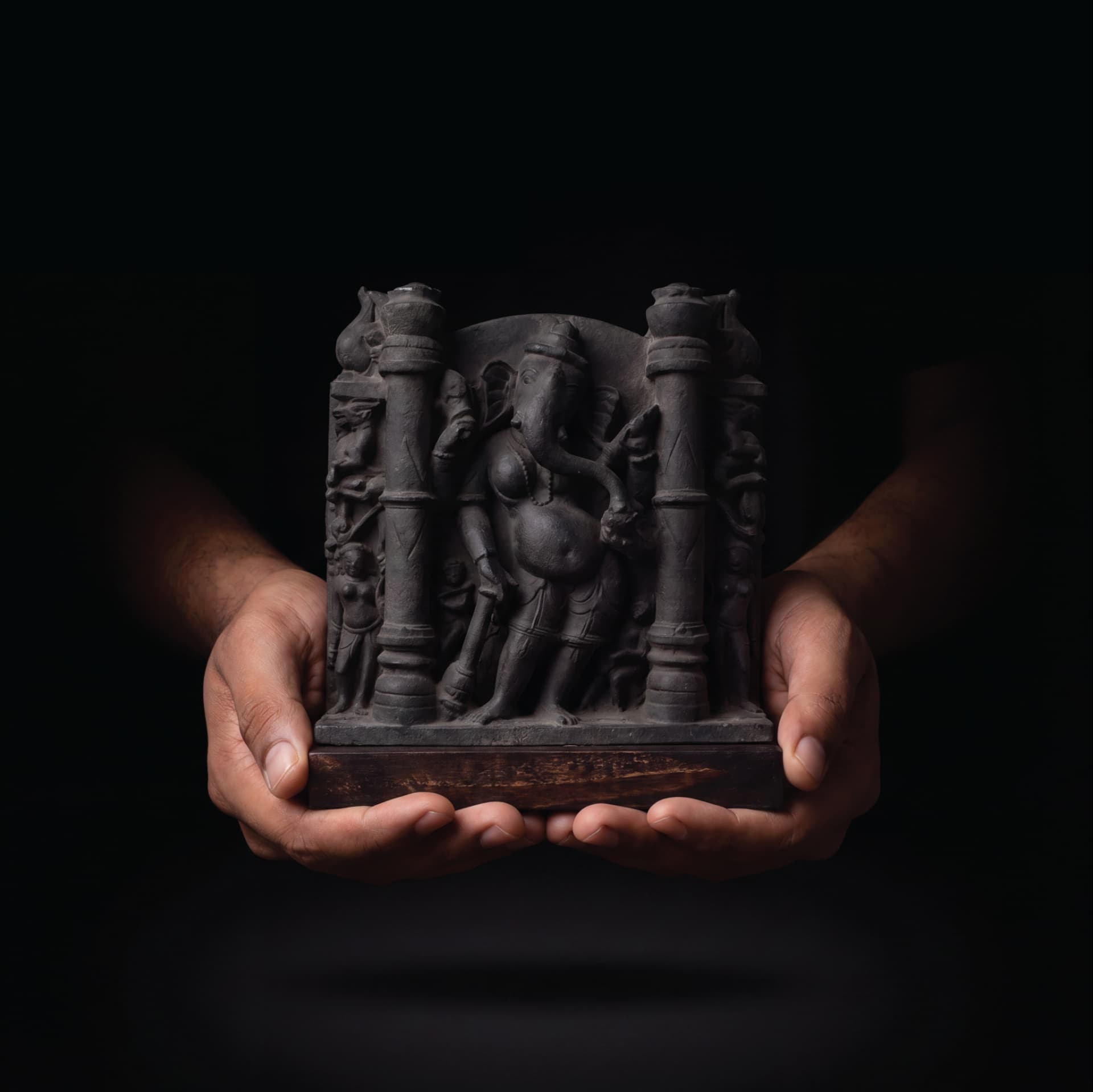 Hands hold a dark stone Vinayaki Sculpture, a detailed museum-grade replica featuring intricate temple art carvings against a black background.