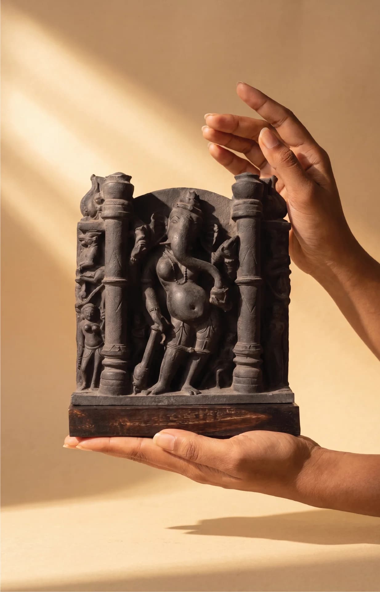 A pair of hands gently holds a dark stone Vinayaki Sculpture, showcasing the intricate 11th-century medieval Indian temple art details.