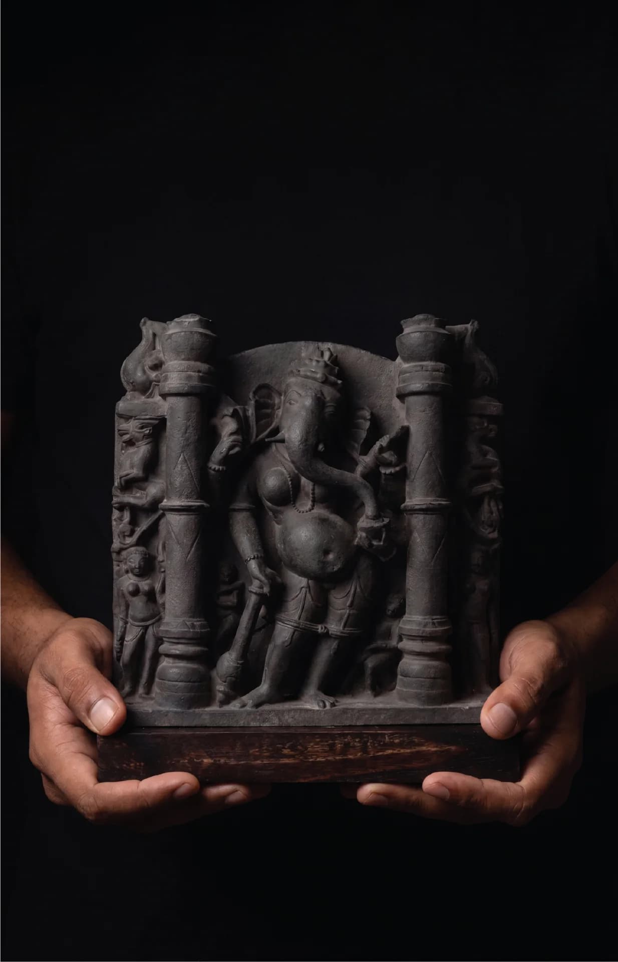 A pair of hands holds a dark stone Vinayaki Sculpture, a detailed museum replica featuring intricate temple iconography.